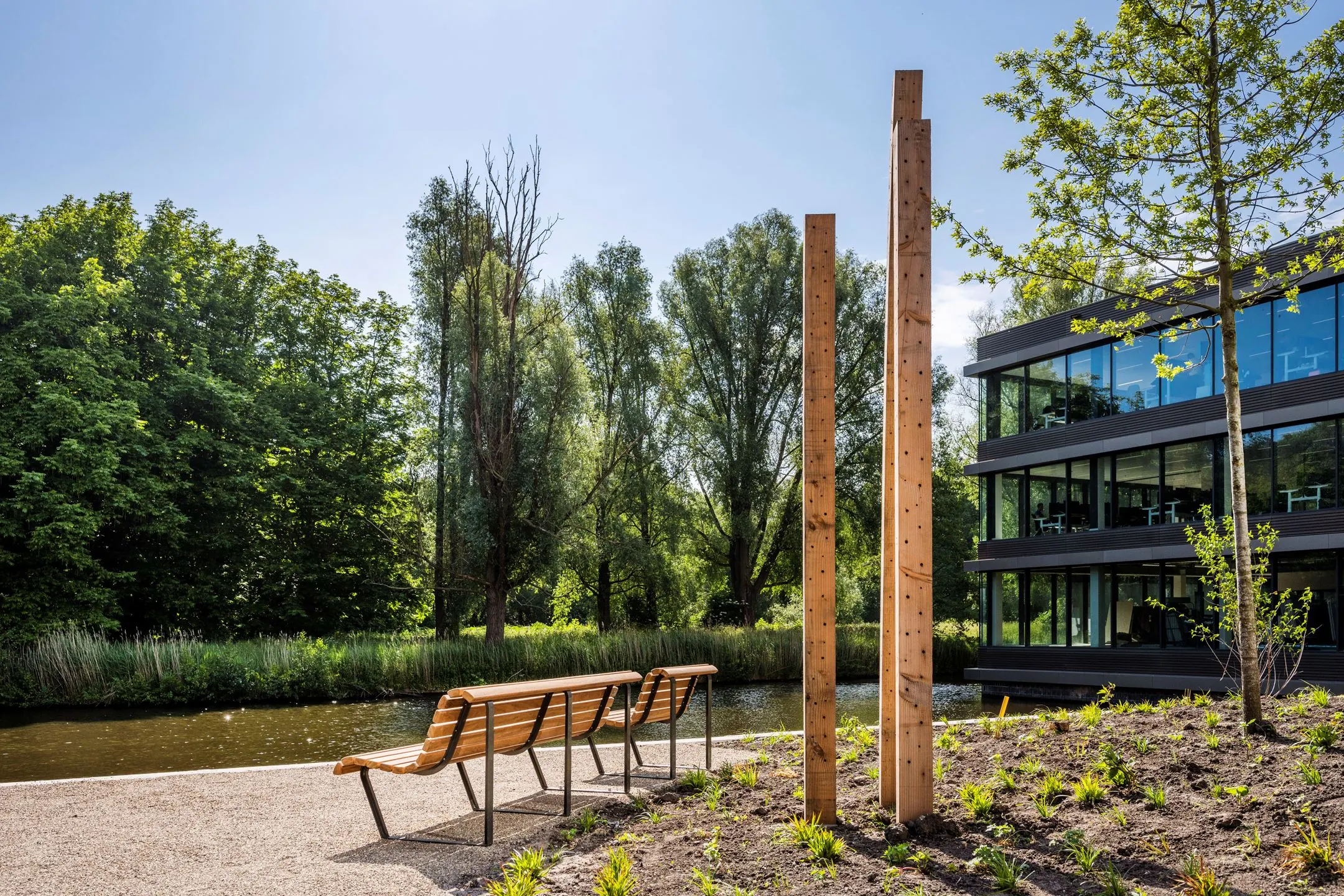 Bankjes aan het water bij een modern kantoorgebouw aan de Nachtwachtlaan, omgeven door bomen en kunstzinnige houten palen.