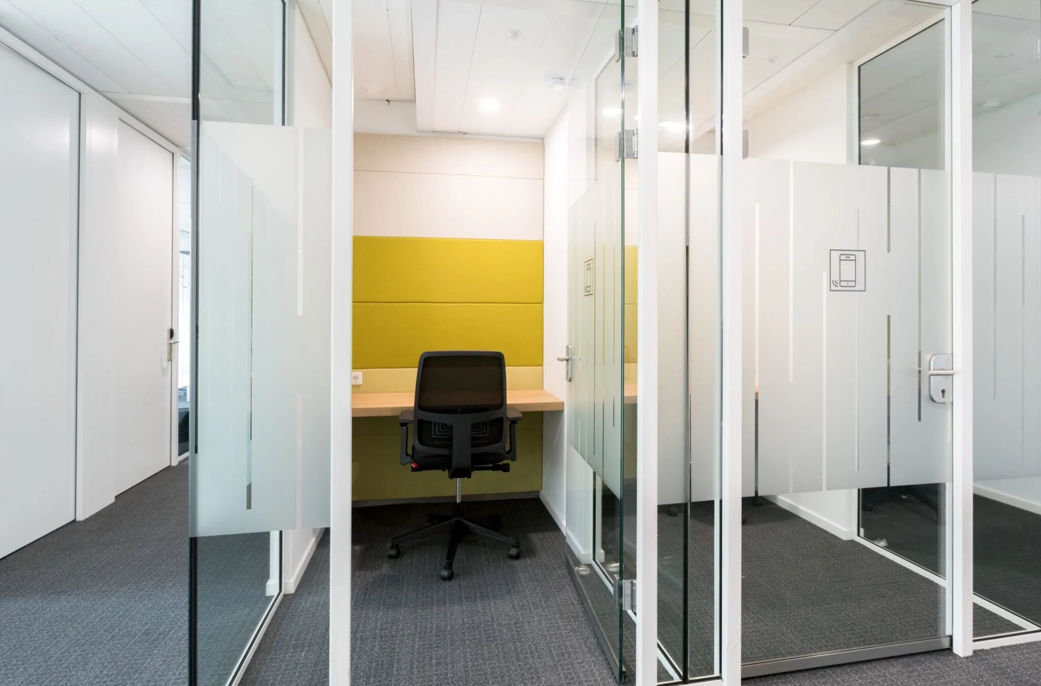 Kleine glazen belruimte met een bureau, bureaustoel en gele wandbekleding in Nevelgaarde 8.