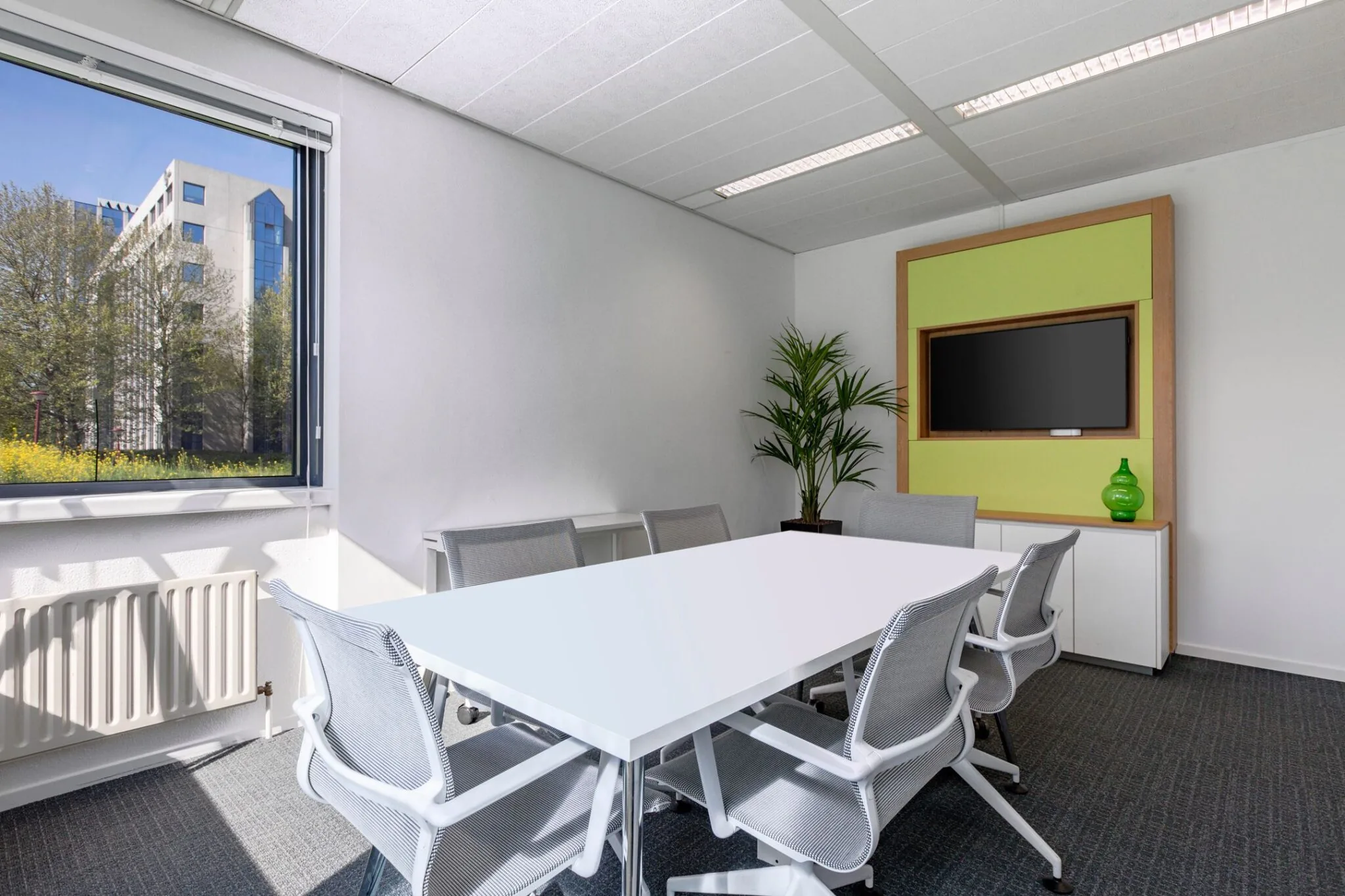 Vergaderruimte in Nevelgaarde 8 met witte tafel, zes stoelen, flatscreen, plant en uitzicht op kantoorpand.
