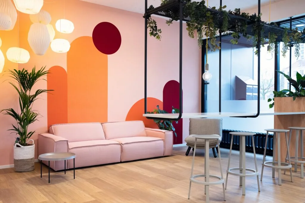 Moderne en kleurrijke zithoek in Newday Arnhem met een zachtroze bank, designverlichting en planten tegen een grafisch beschilderde muur.