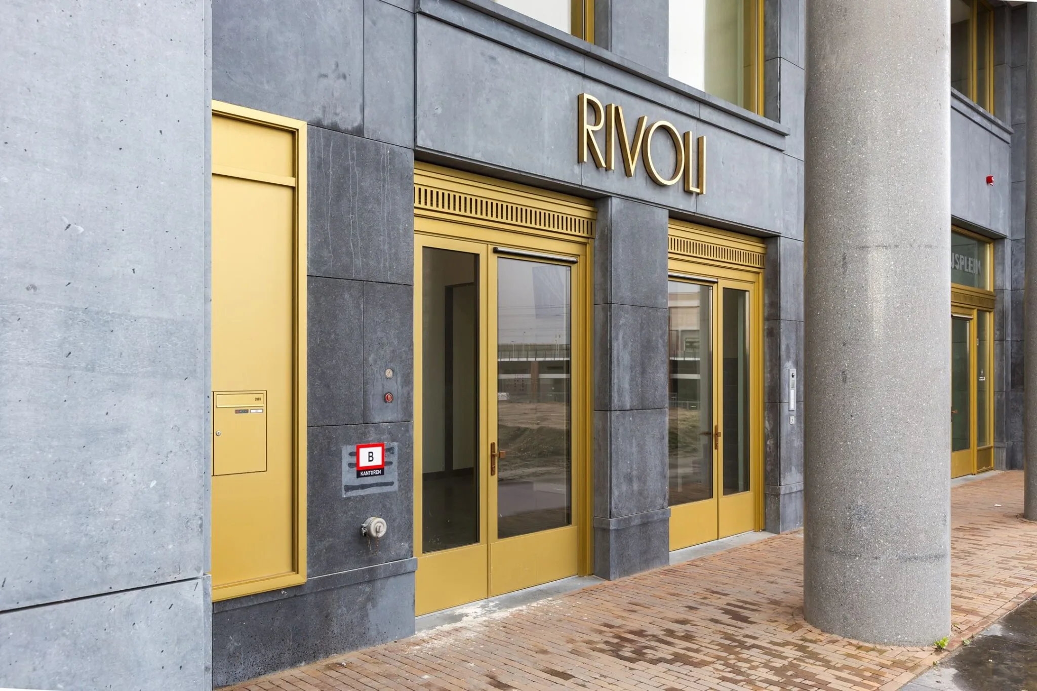 Vooraanzicht van de ingang van het kantoorgebouw Rivoli aan de Parijsboulevard 209 met goudkleurige deuren en grijze gevel.