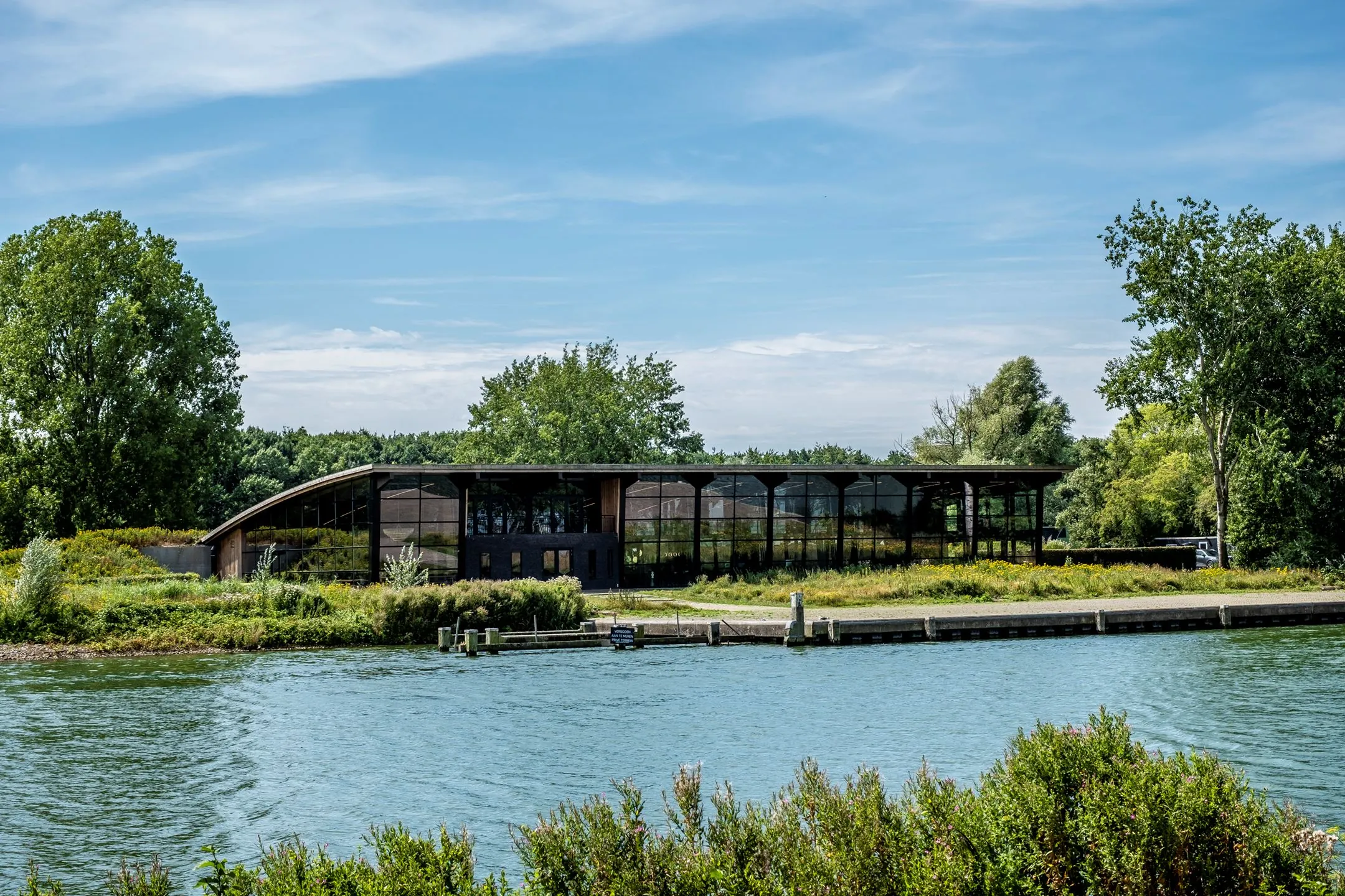Modern gebouw aan het water met grote glazen gevel bij Plaatweg 15, omringd door groen.