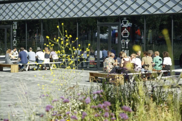 Groep mensen zit buiten op banken te lunchen bij een modern horecapand, omringd door wilde bloemen.