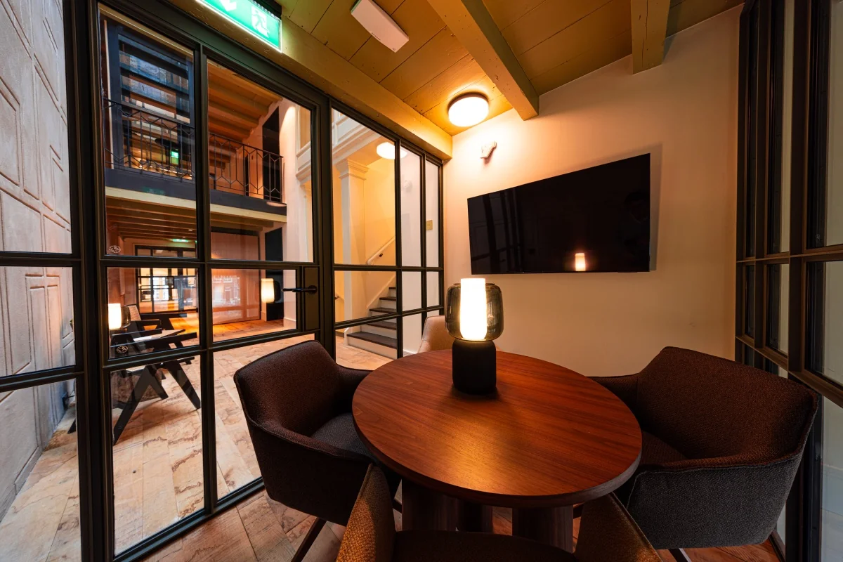 Een moderne vergaderruimte met een ronde tafel, stoelen en een lamp, gezien door glazen panelen, met een open ruimte op de achtergrond.