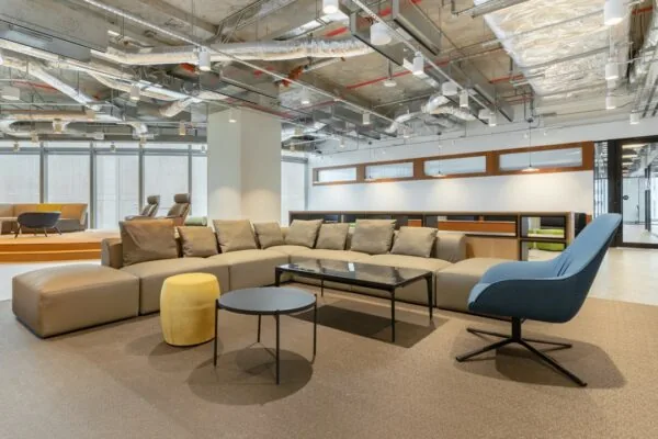 Moderne loungehoek met een grote grijze hoekbank, kleurrijke stoelen en industriële plafondafwerking in een kantoorruimte op Rotterdam Airportplein 22.