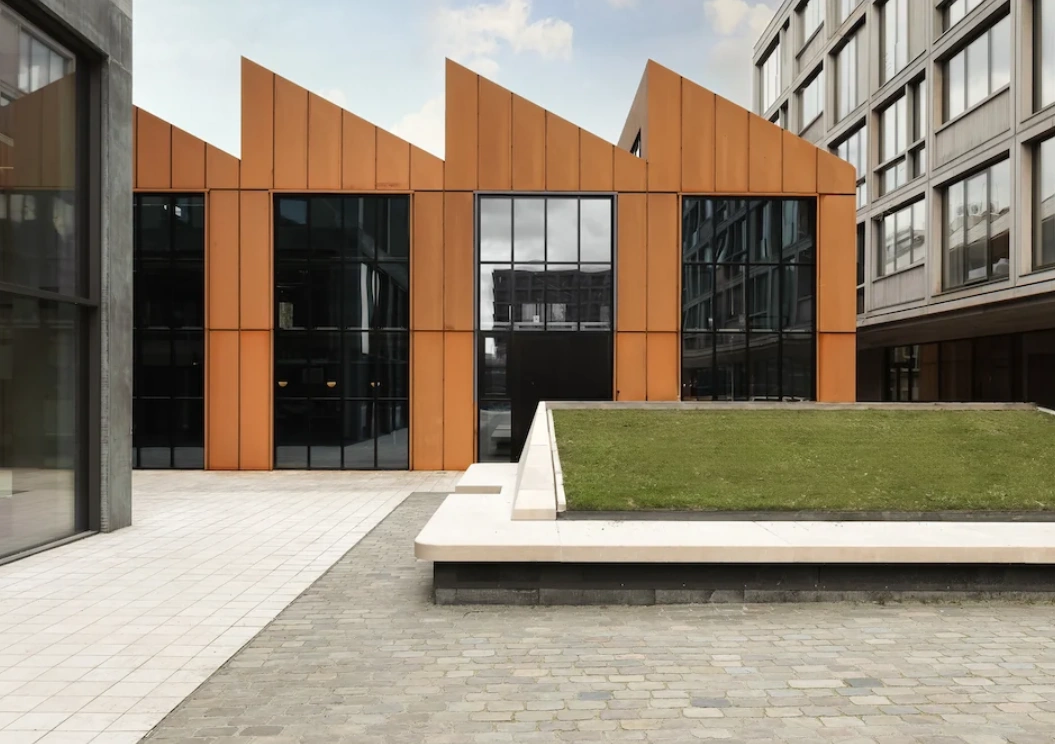 Een modern gebouw met oranje gevelbekleding en grote ramen naast een geplaveide binnenplaats en groengebied.