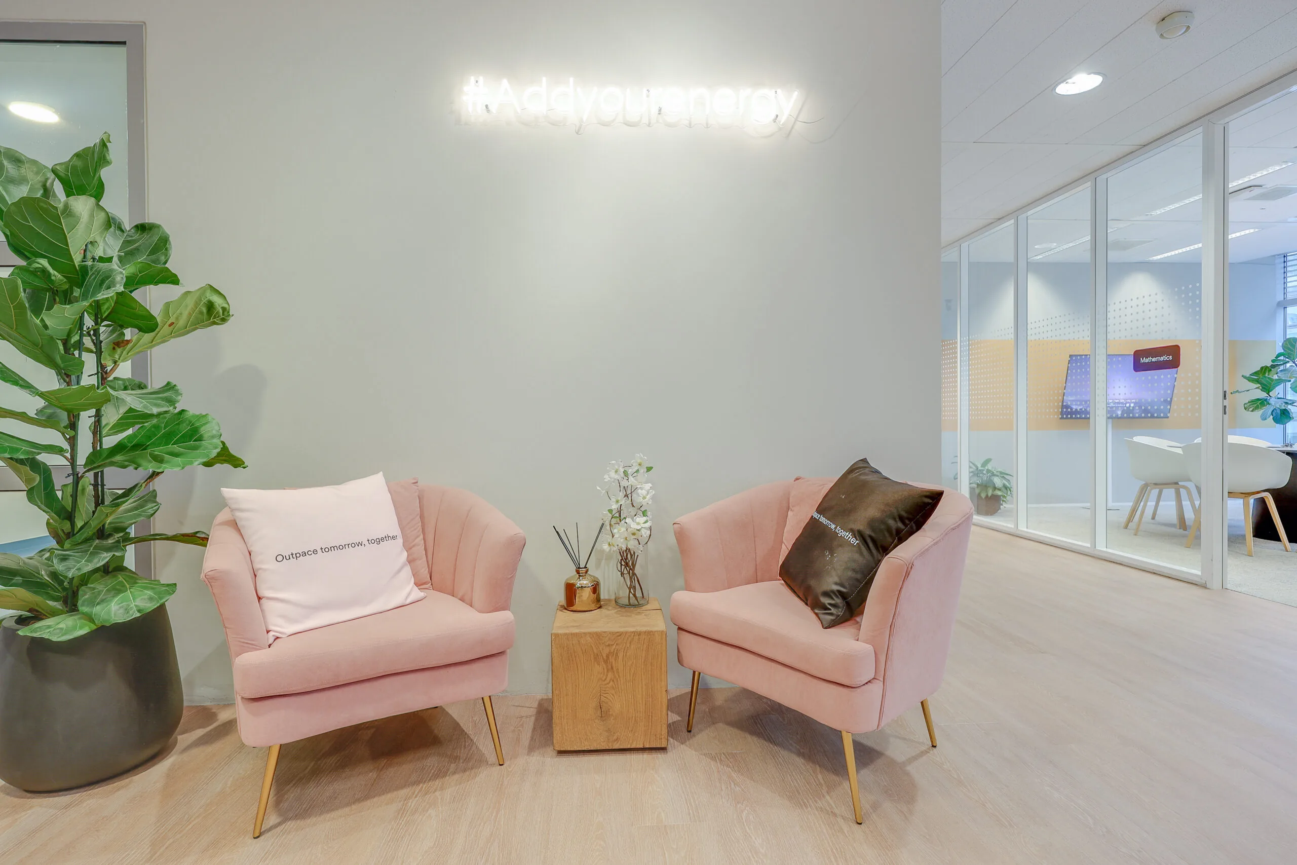 Twee roze stoelen met kussens staan naast een plant in een moderne ruimte met een #Addyourenergy lichtreclame.