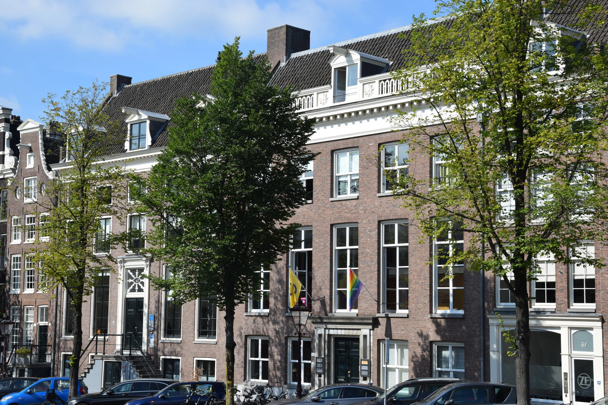 Vooraanzicht van een klassiek Amsterdams grachtenpand met regenboog- en gele vlag bij de ingang.