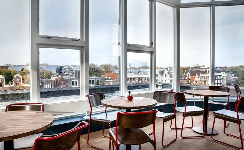 Moderne zithoek met houten tafels en stoelen bij grote ramen met uitzicht op Amsterdam vanaf Weesperstraat 61-105.