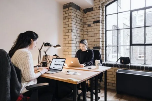 Twee vrouwen werken op laptops in een modern kantoor aan de Weesperstraat met bakstenen muren en grote ramen.