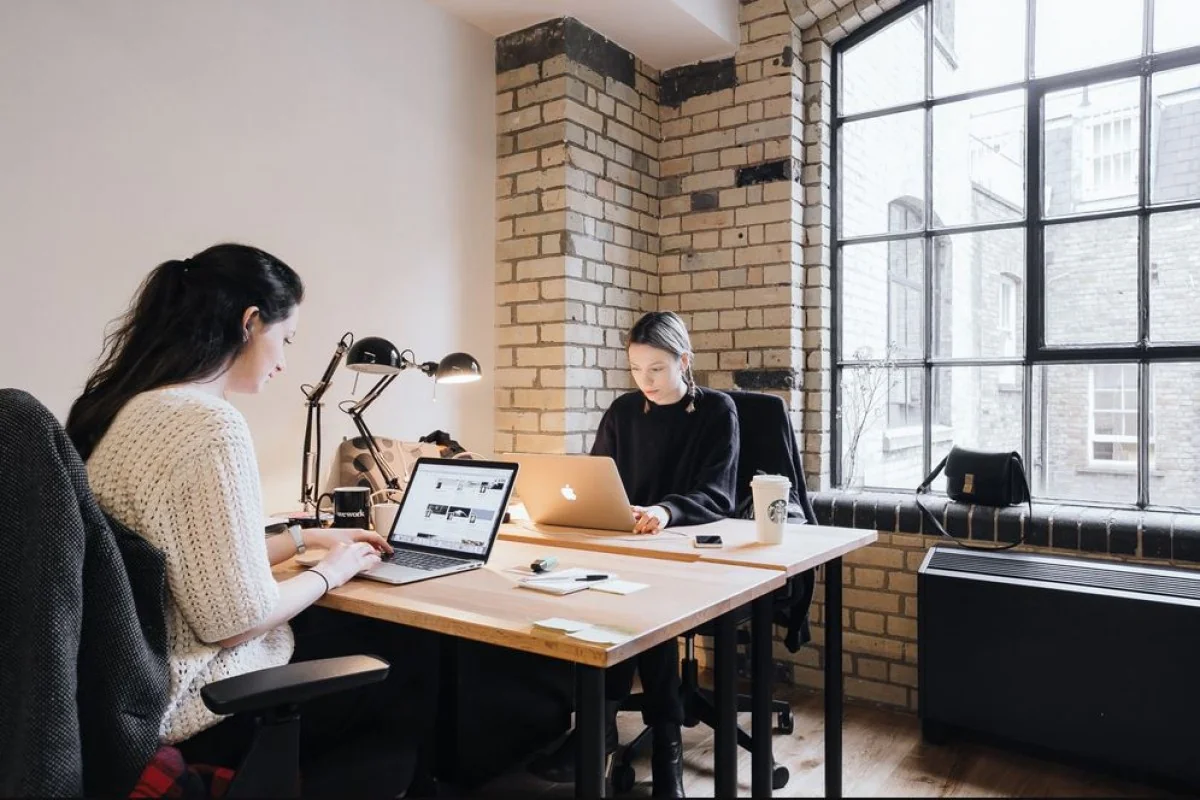 Twee vrouwen werken op laptops in een modern kantoor aan de Weesperstraat met bakstenen muren en grote ramen.