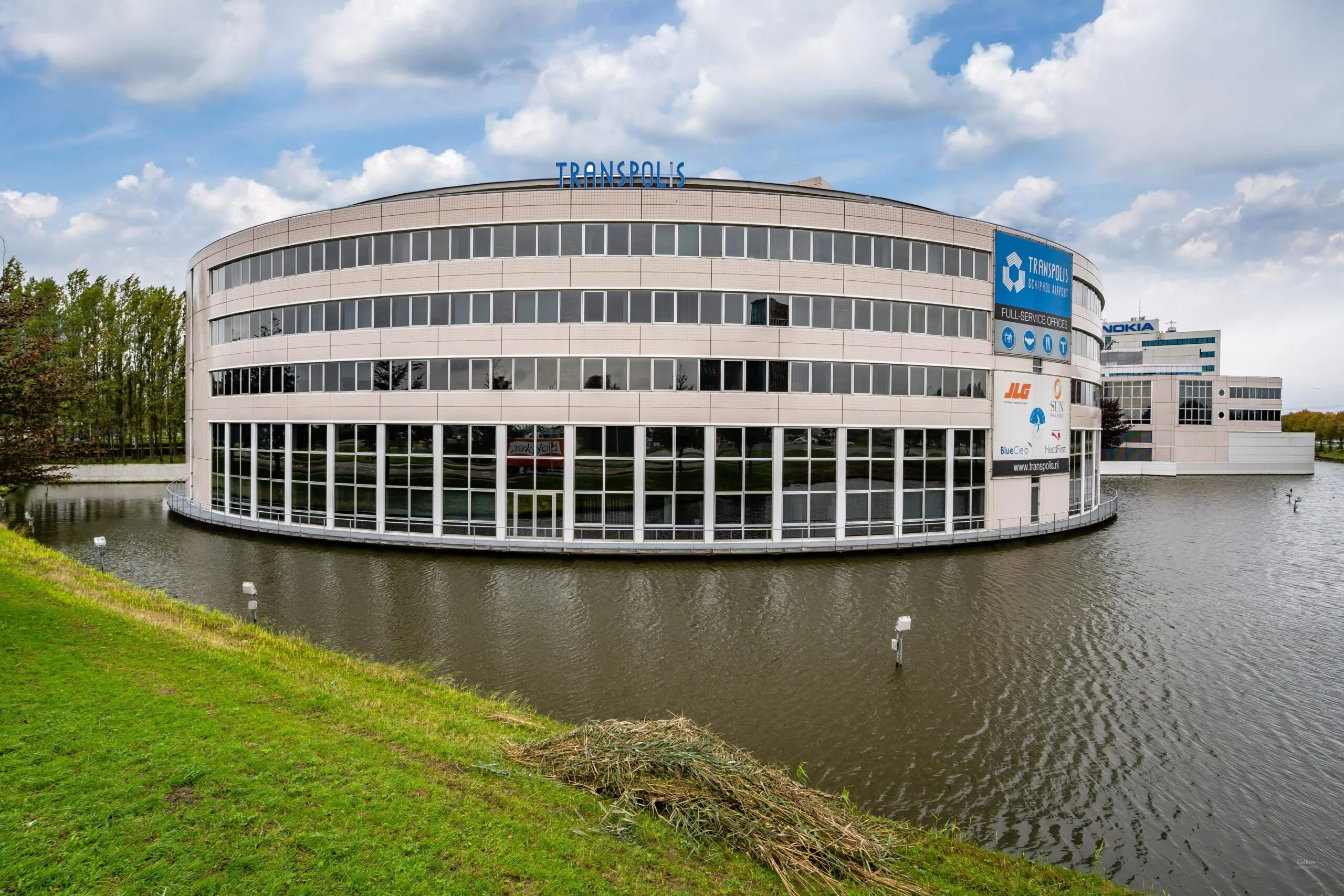 Een modern kantoorgebouw genaamd Transpolis, omgeven door water en een grasveld.