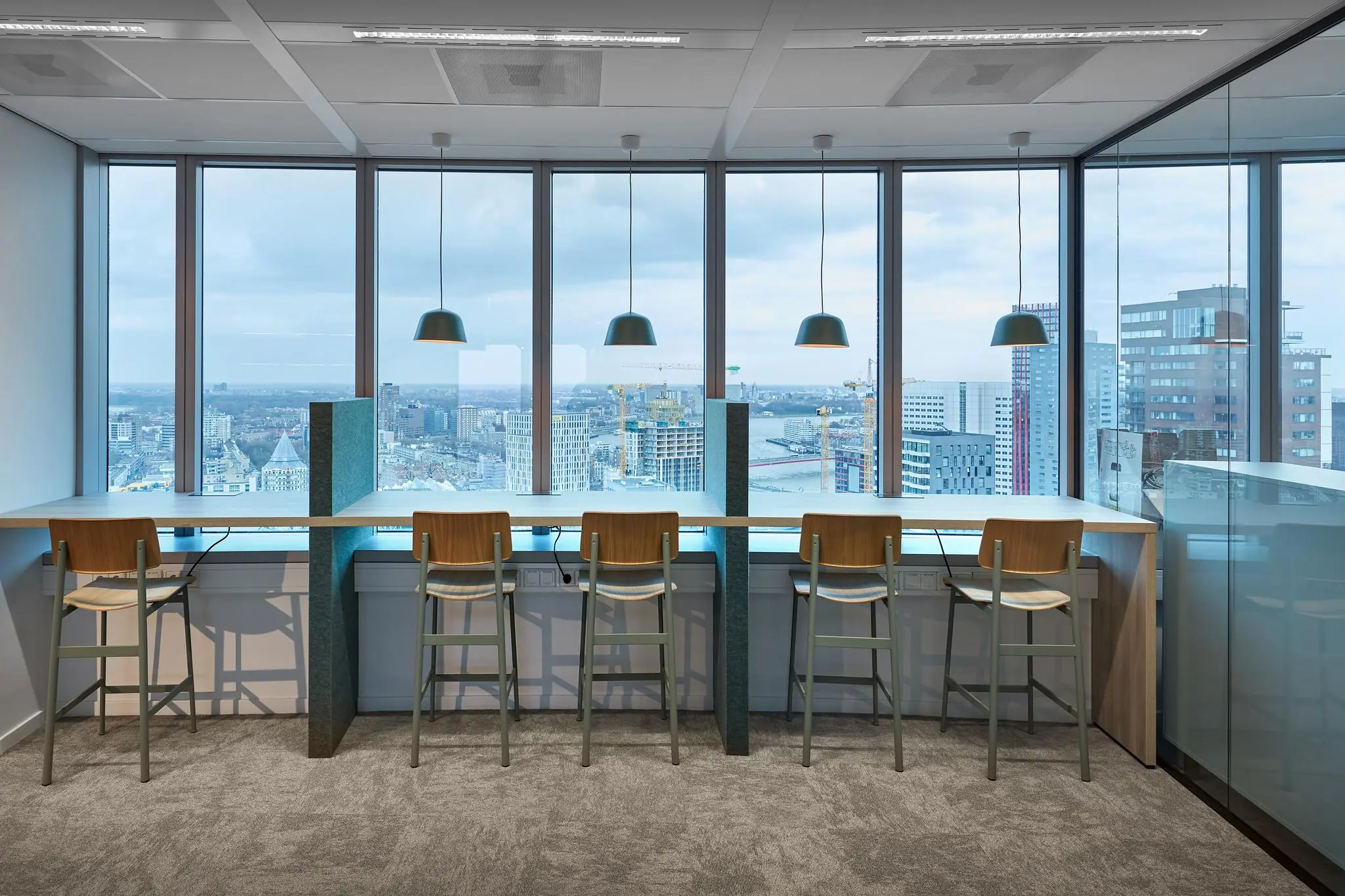 Moderne werkplekken met hoge stoelen en hanglampen bij het raam op een hoge verdieping van Blaak 555, met uitzicht over de skyline van Rotterdam.