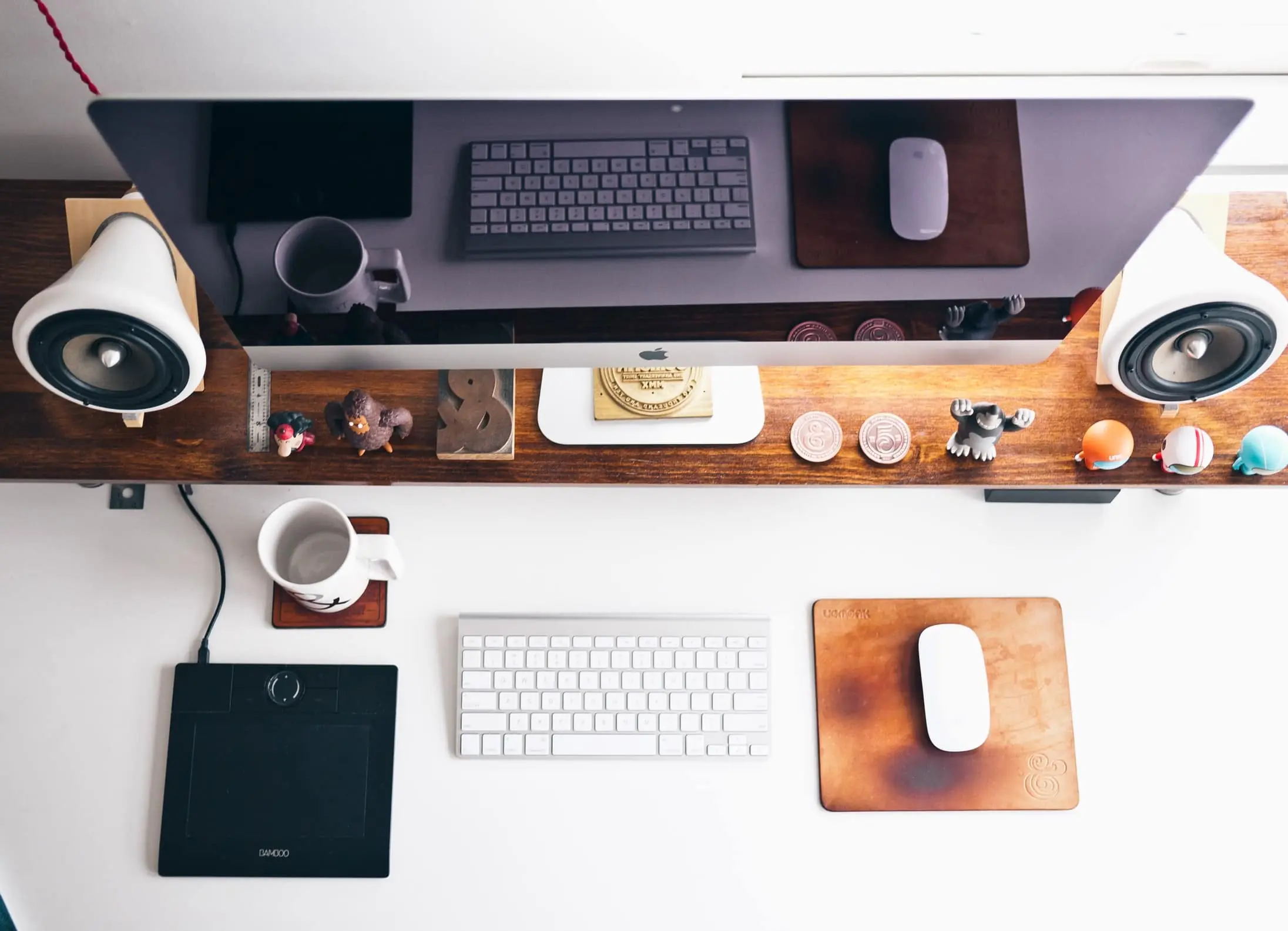 Minimalistisch bureau met computer, toetsenbord, tekentablet en diverse bureau-gadgets en figuurtjes op een houten plank.