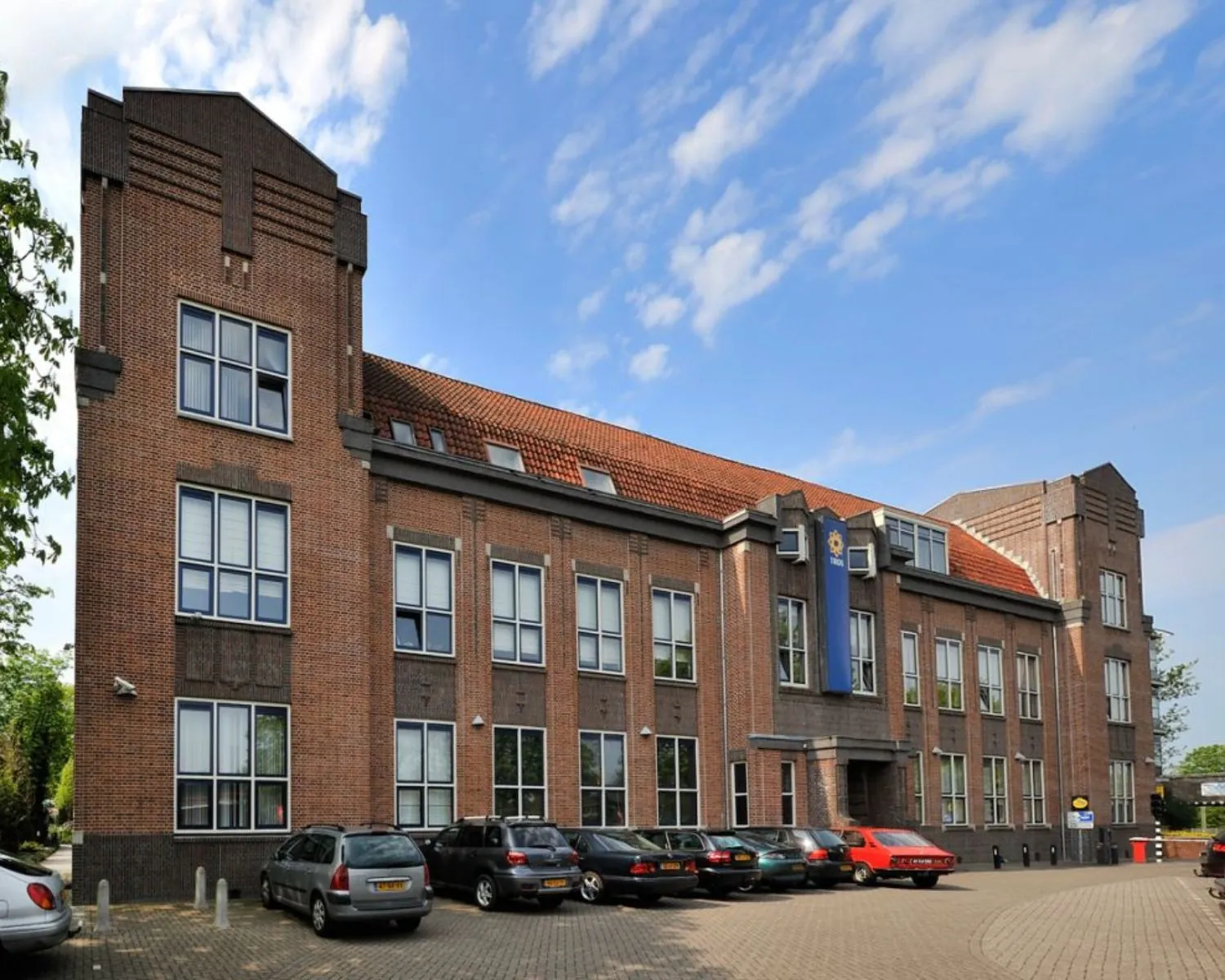 Bakstenen politiebureau met rood dak en geparkeerde auto's op de voorgrond, onder een deels bewolkte hemel.