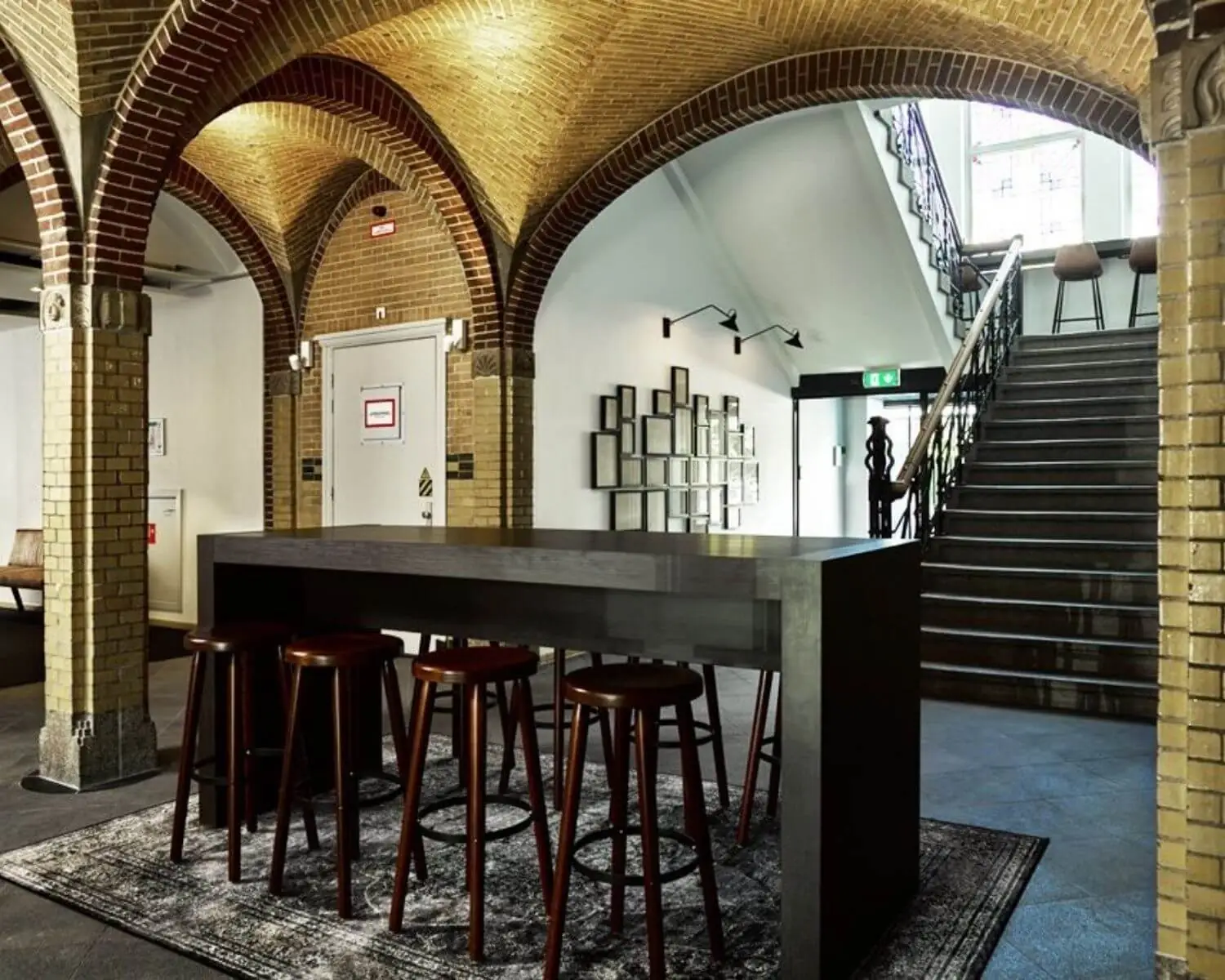 Modern ingerichte ruimte met hoge tafel en barkrukken onder bakstenen gewelven naast een trap.
