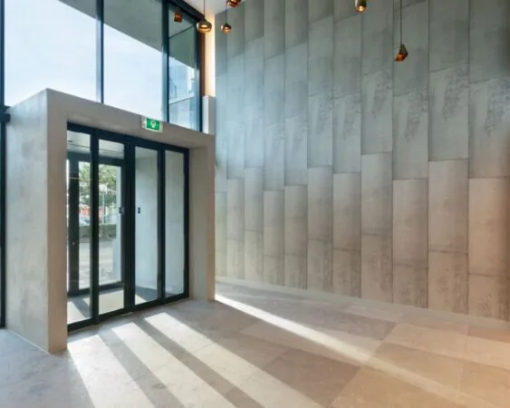 Moderne entreehal met glazen schuifdeur, hoge plafonds en betonnen wandpanelen.