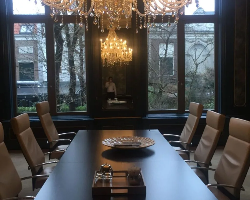 Luxe vergaderruimte met leren stoelen, kristallen kroonluchters en grote ramen met uitzicht op een tuin.