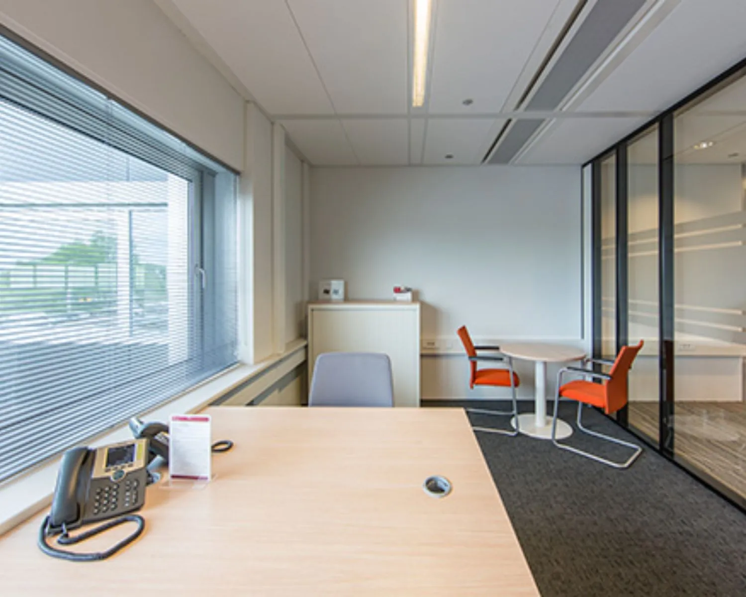 Modern ingericht kantoor met een bureau, vergadertafel en twee oranje stoelen bij een glazen wand.