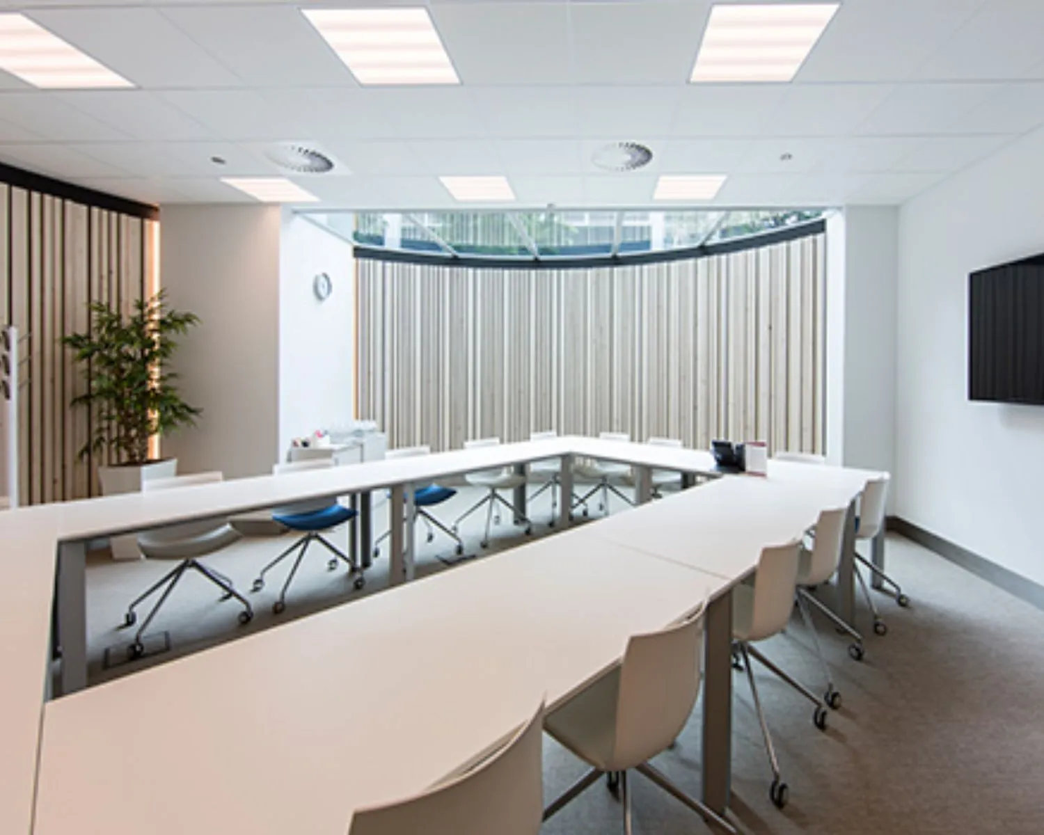 Moderne vergaderruimte met U-vormige opstelling van witte tafels en stoelen, grote ramen en een flatscreen aan de muur.