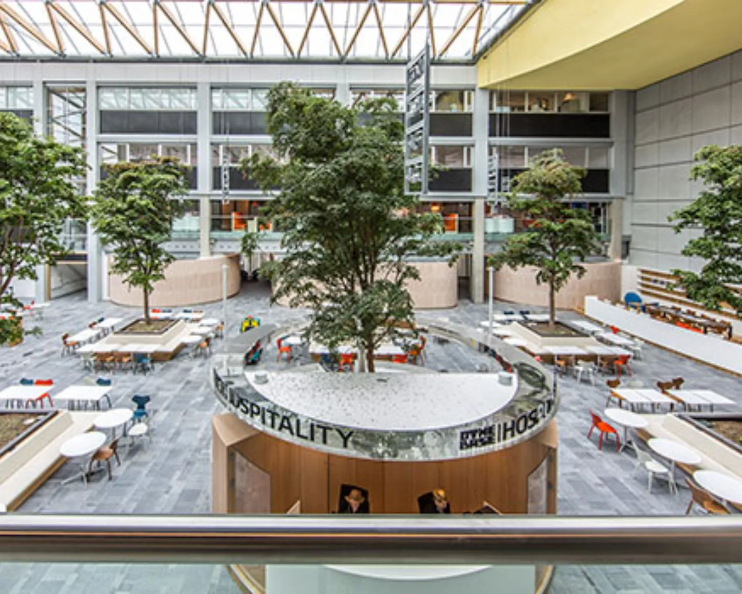Moderne, open binnentuin met bomen, zitplaatsen en een ronde balie met de tekst "HOSPITALITY" in een kantoorgebouw.