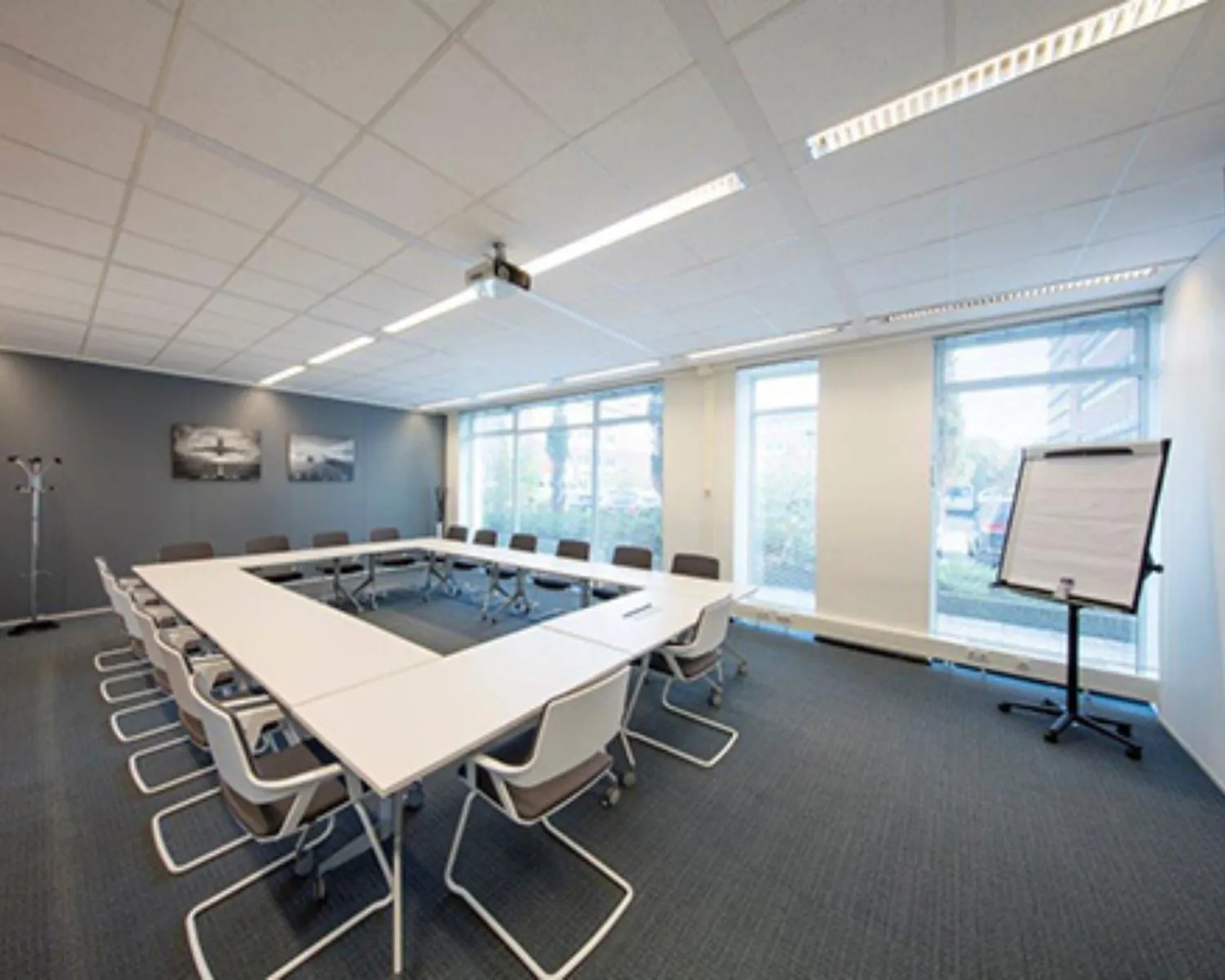 Moderne vergaderruimte met U-vormige opstelling, conferentiestoelen, flipover en grote ramen.
