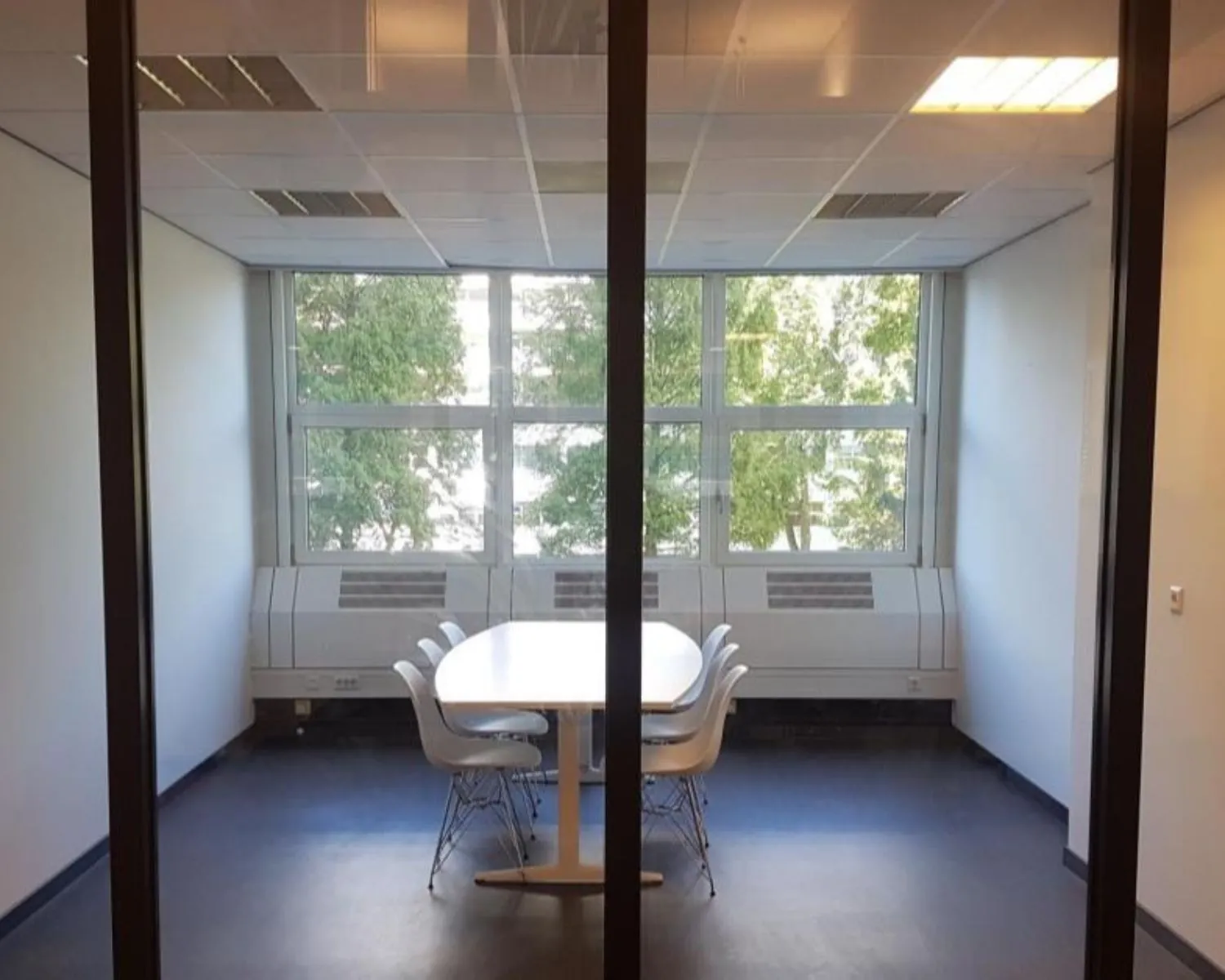 Vergaderruimte met een witte tafel en zes stoelen, gelegen aan een raam met uitzicht op bomen.