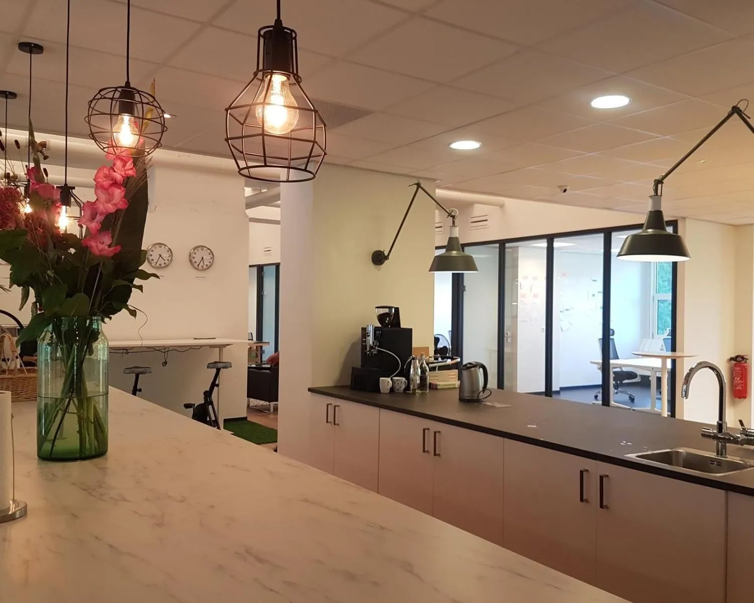 Moderne kantoorkeuken met hanglampen, bloemen op het aanrecht en zicht op vergaderruimtes.