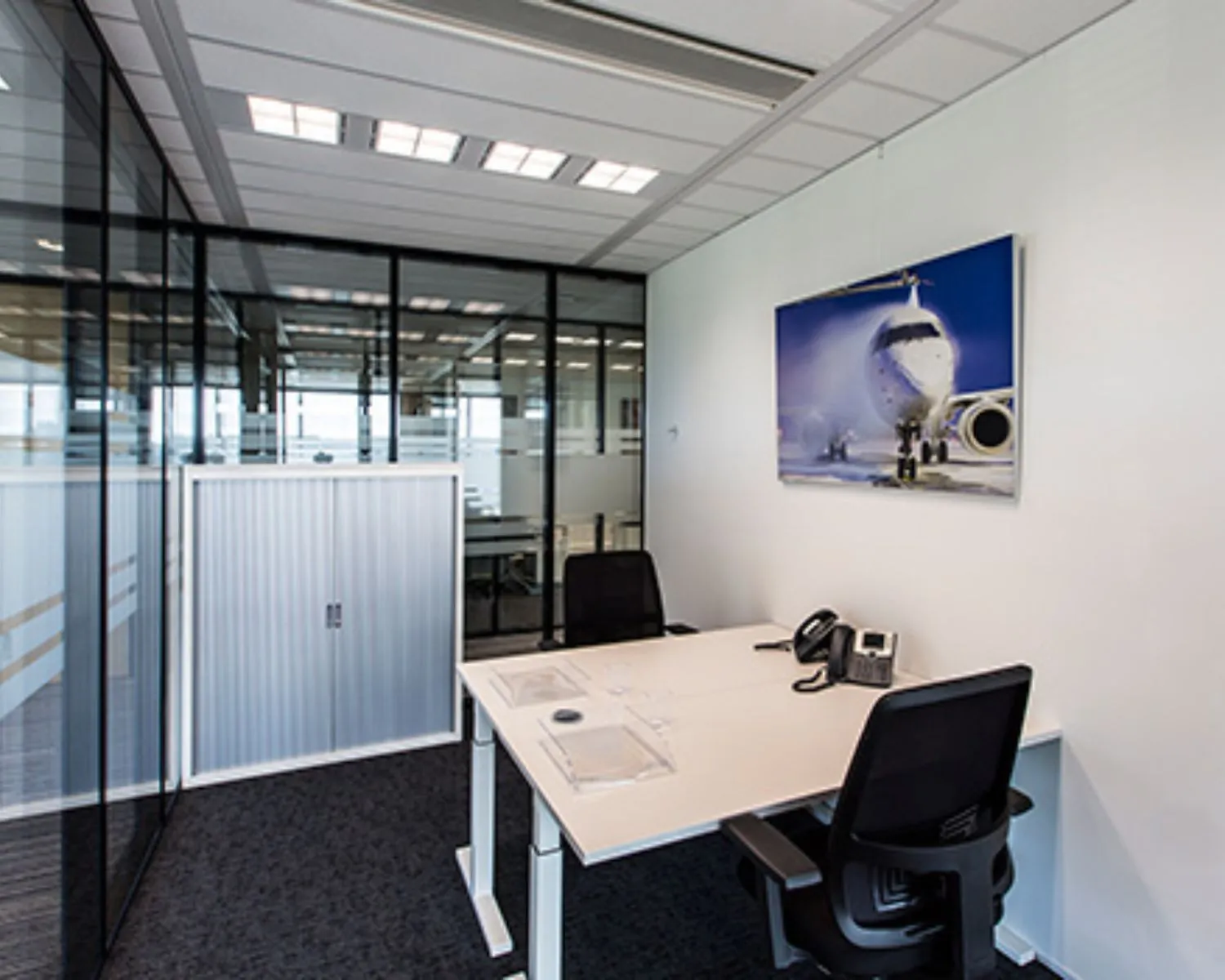 Modern kantoor met glazen wanden, twee bureaustoelen en een wandfoto van een vliegtuig.