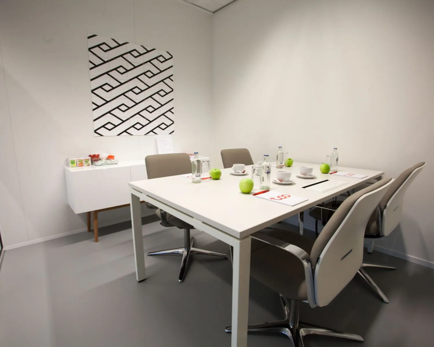Vergaderruimte met een witte tafel, zes stoelen, groene appels, kopjes en notitieblokken, en een modern kunstwerk aan de muur.