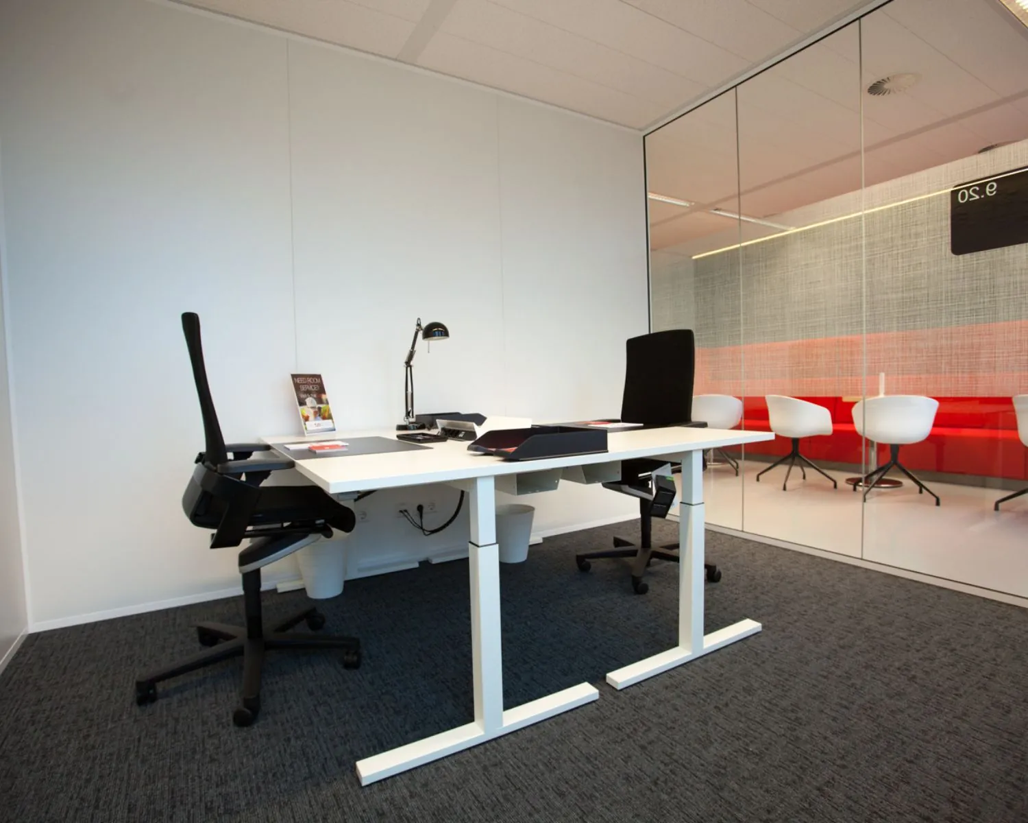 Modern kantoor met twee bureaustoelen, een wit bureau en glazen wand met zicht op een vergaderruimte.