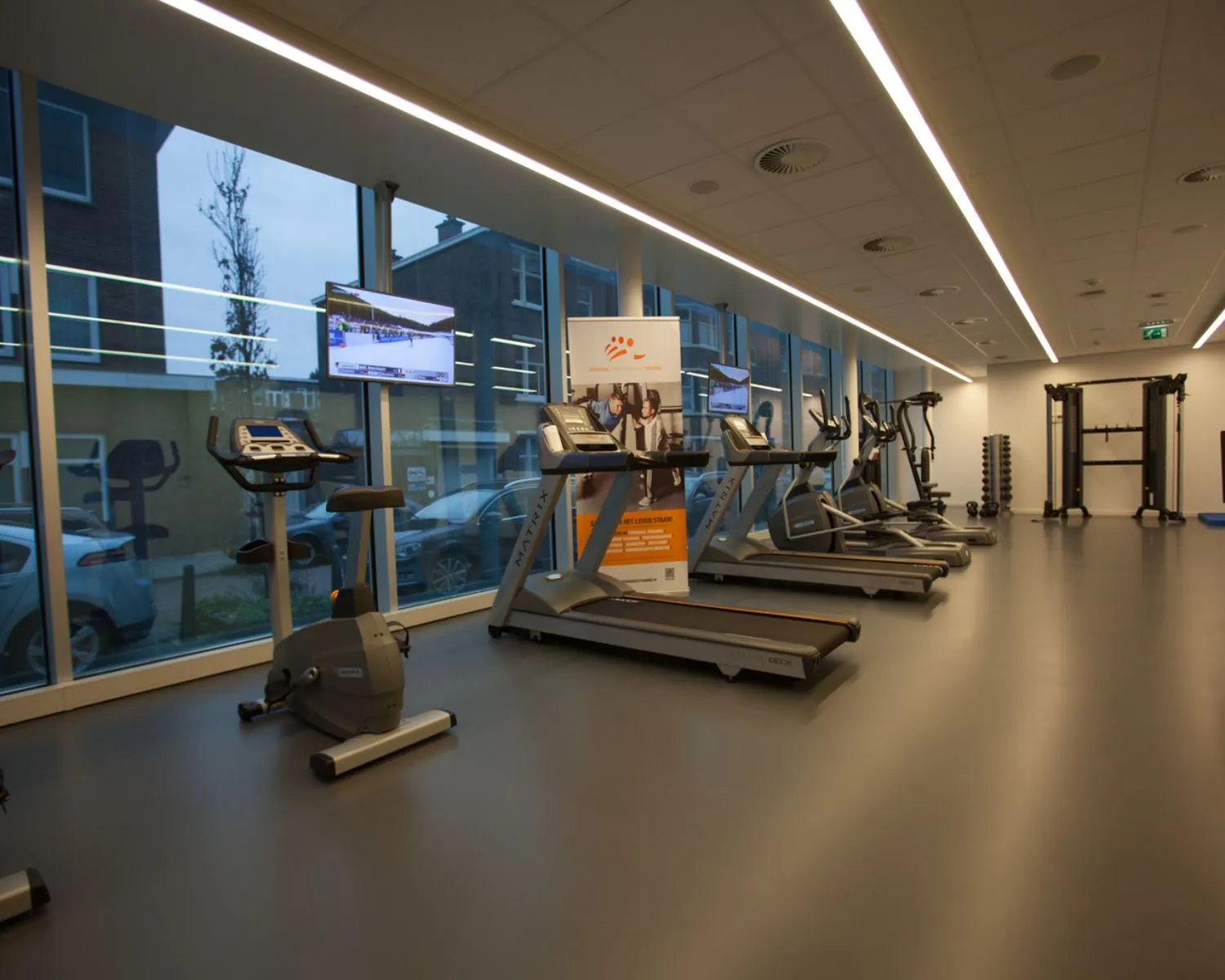 Moderne fitnessruimte met cardioapparatuur, televisieschermen en grote ramen met uitzicht op de straat.