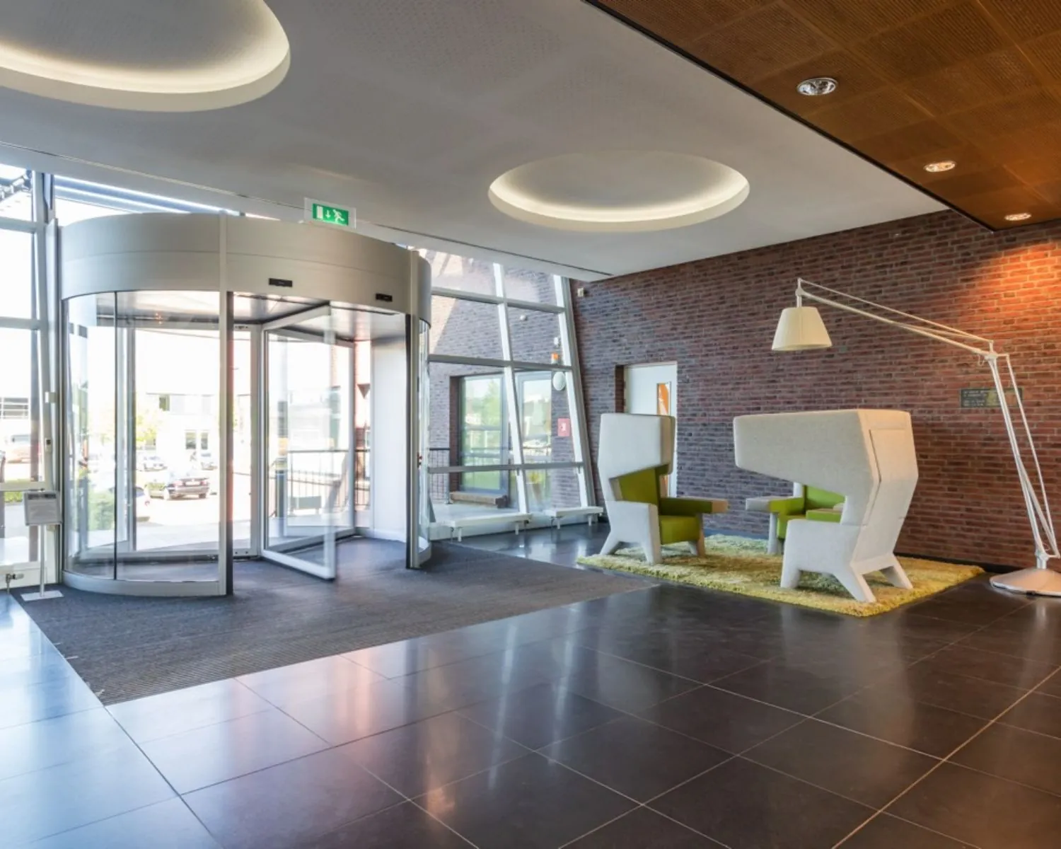 Moderne entreehal met draaideur, designstoelen en een grote vloerlamp tegen een bakstenen muur.