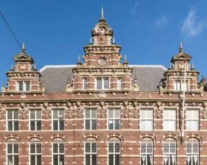 Voorgevel van een monumentaal, rijk versierd bakstenen pand met klassieke ornamenten en trapgevels tegen een blauwe lucht.