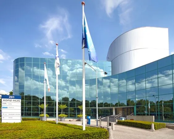 Moderne kantoorgebouw met glazen gevel van het ADFIZ-Huis, met vlaggen en een naambord in de voortuin.