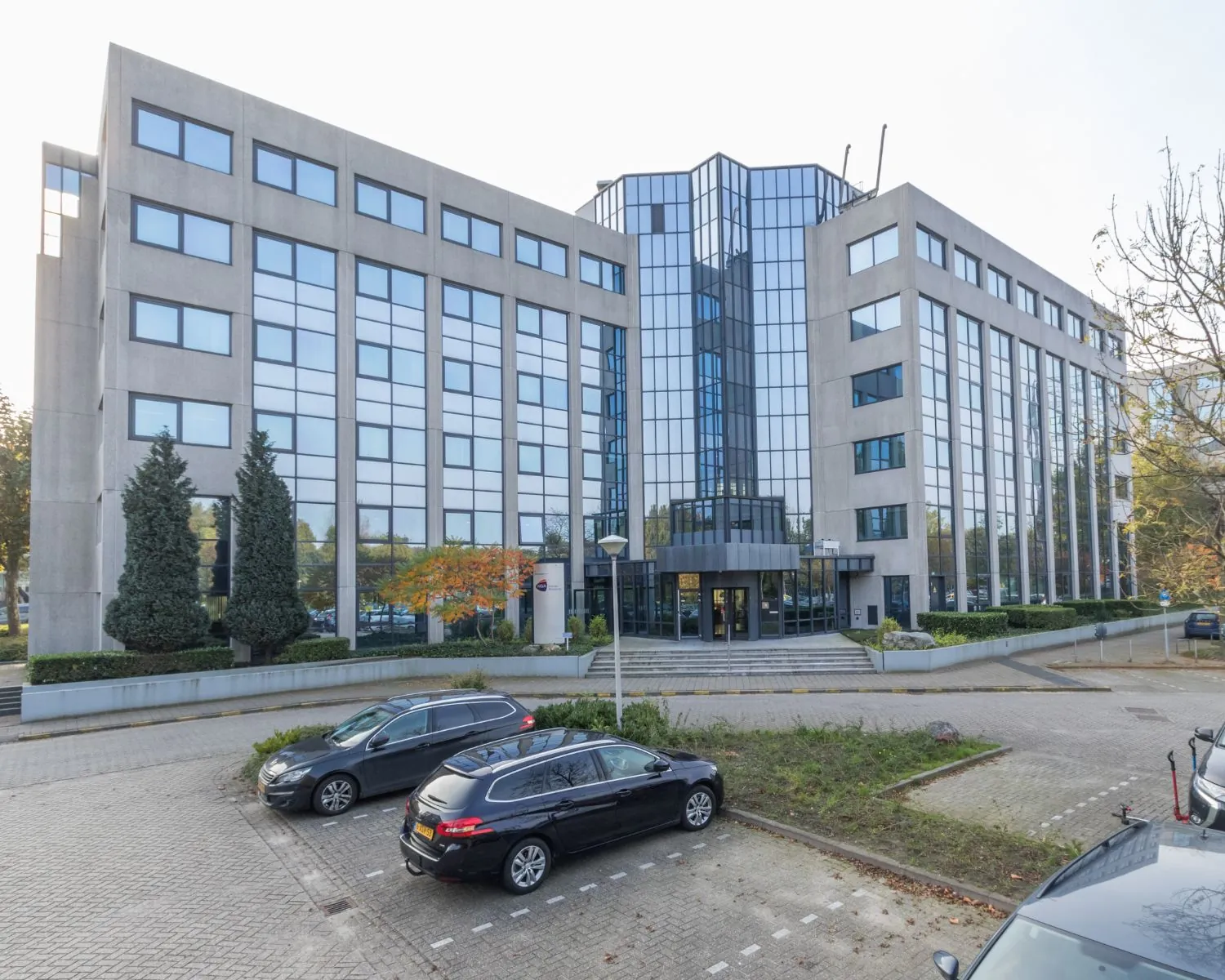 Moderne kantoorgebouw met spiegelende ramen en een parkeerplaats ervoor.