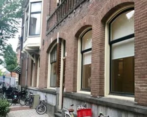 Bakstenen gevel van een oud pand met hoge ramen en geparkeerde fietsen op de stoep.