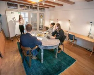 Vier mensen hebben een vergadering in een sfeervolle kantoorruimte terwijl een vrouw iets uitlegt op een whiteboard.