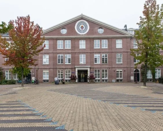 Vooraanzicht van een klassiek bakstenen schoolgebouw met symmetrische ramen en een ronde raampartij in de topgevel, geflankeerd door herfstkleurige bomen.