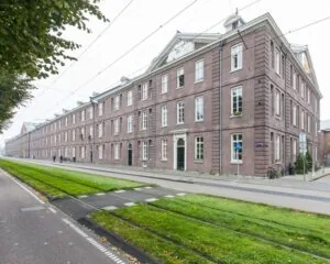 Langgerekt historisch bakstenen gebouw met witte kozijnen aan een straat met trambaan in een stedelijke omgeving.