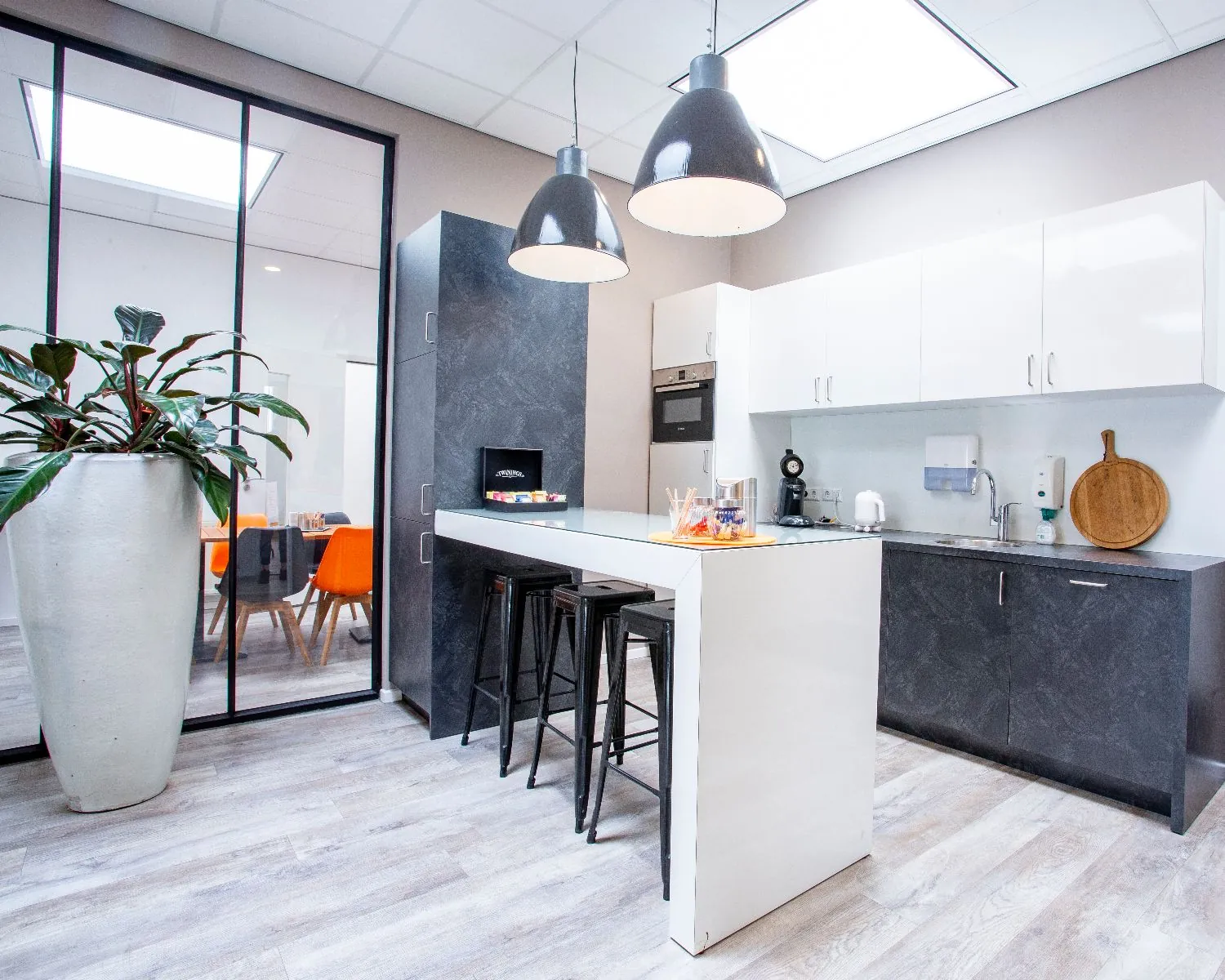 Moderne kantoorkeuken met een kookeiland, zwarte barkrukken, hanglampen en zicht op een vergaderruimte met oranje stoelen.