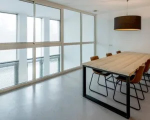 Vergaderruimte met een grote houten tafel, acht bruine stoelen en een zwarte hanglamp, omgeven door glazen wanden.