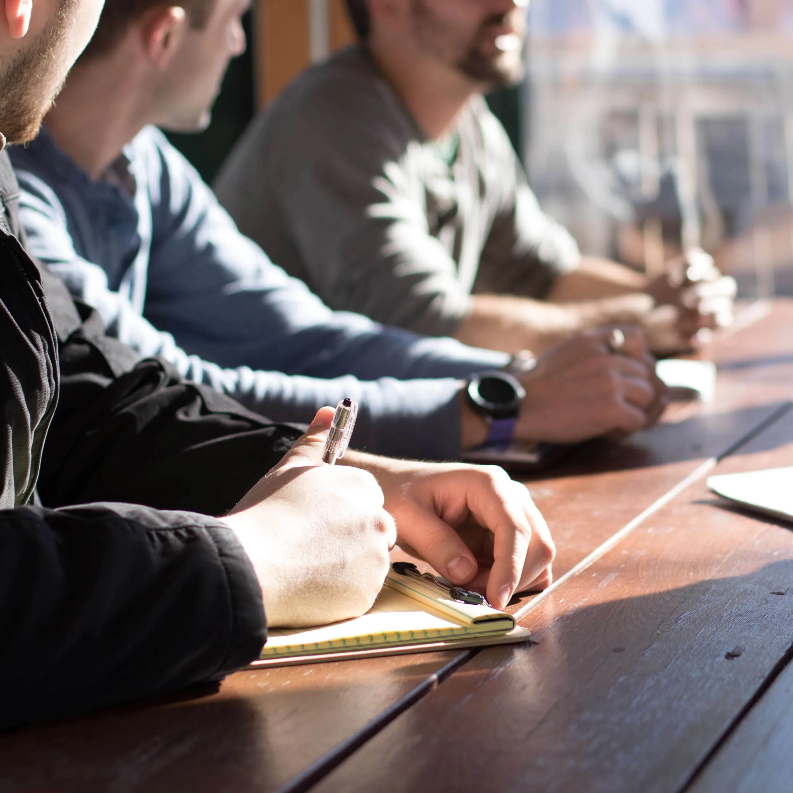 Drie mannen zitten aan een houten vergadertafel terwijl één van hen aantekeningen maakt in een notitieblok.