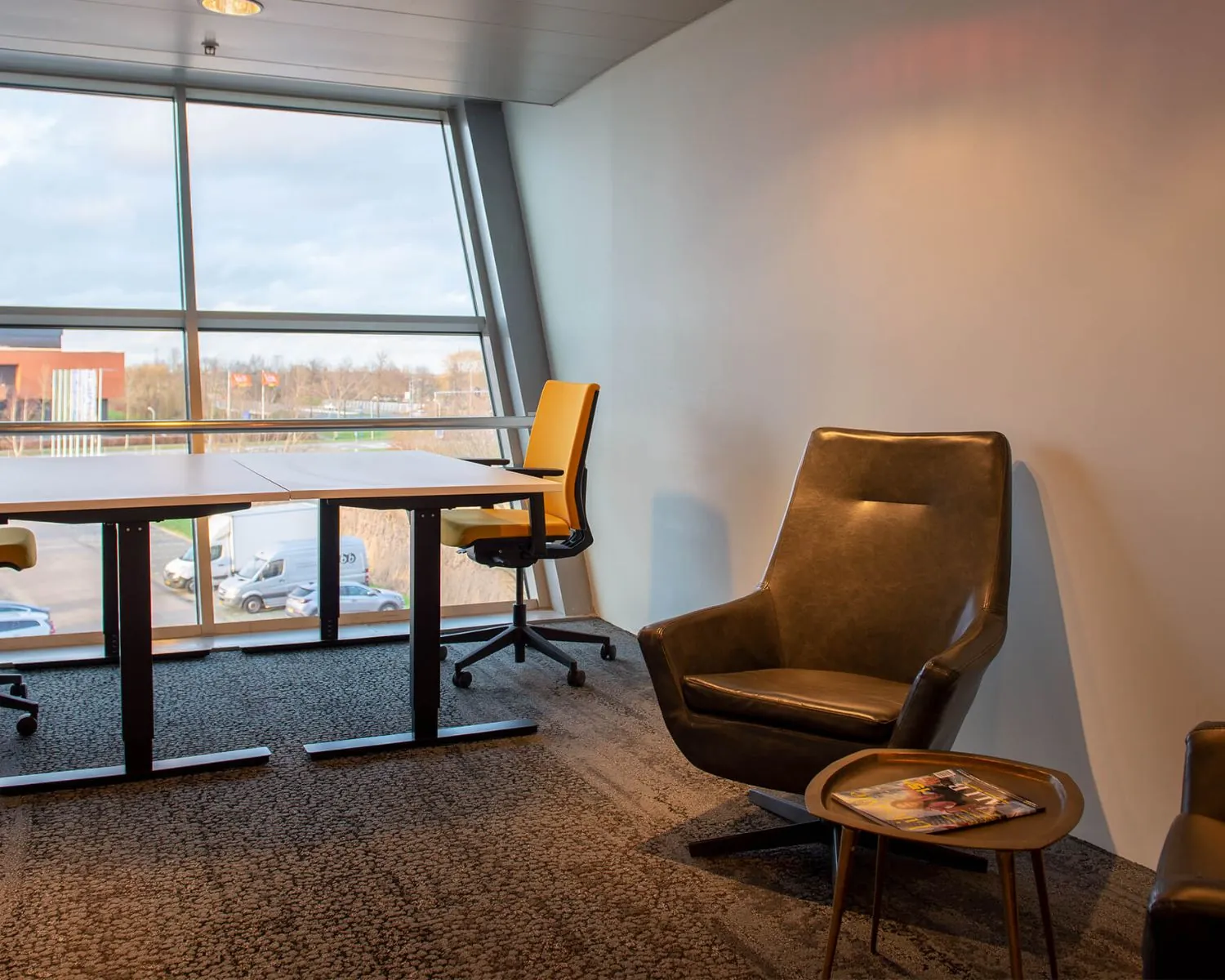 Modern kantoorinterieur met grote ramen, gele bureaustoelen, een leren fauteuil en een bijzettafeltje met tijdschriften.