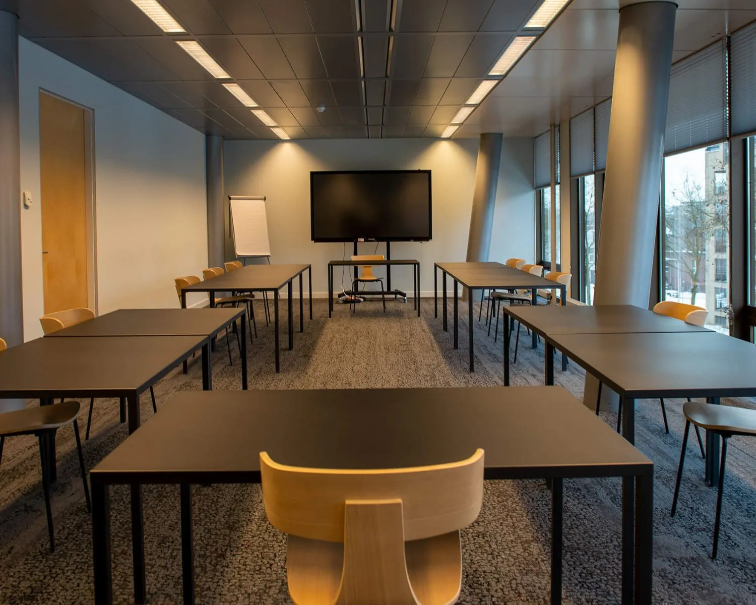 Vergaderruimte met rechte opstelling van tafels en stoelen, een scherm en een flip-over aan de voorzijde.