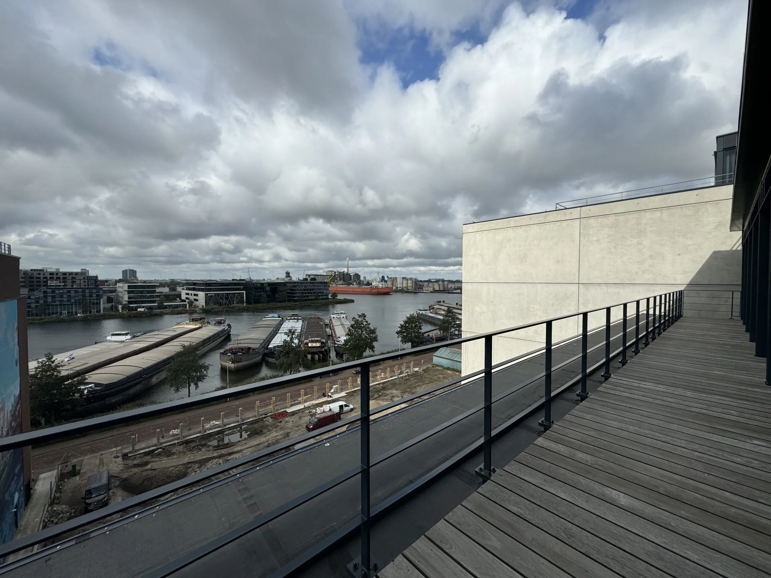 Uitzicht vanaf een houten terras met metalen hek op een rivier met aangemeerde boten onder een bewolkte lucht.