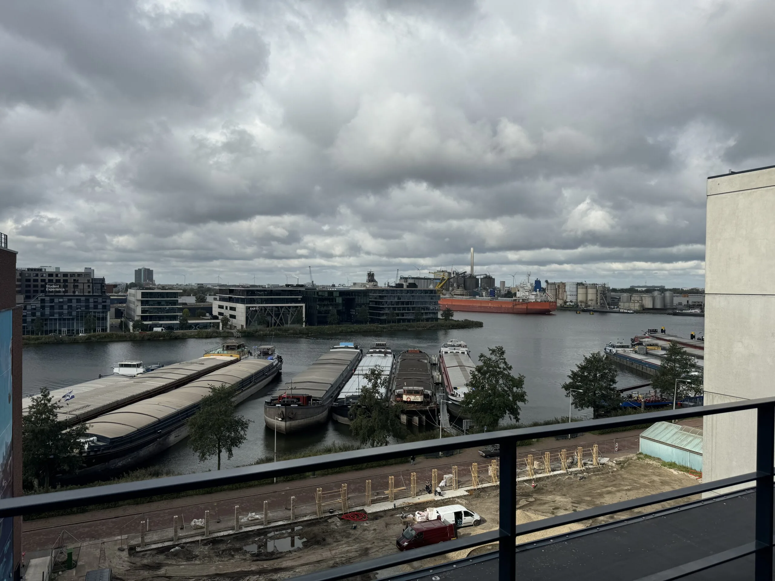 Uitzicht op een rivier met aangemeerde vrachtschepen en industriële gebouwen aan de overkant onder een bewolkte hemel.