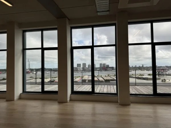Uitzicht op een haven en stedelijke skyline door grote ramen in een modern gebouw.
