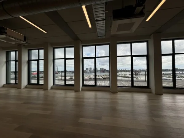 Een leeg interieur met grote ramen die uitkijken op een havengebied.