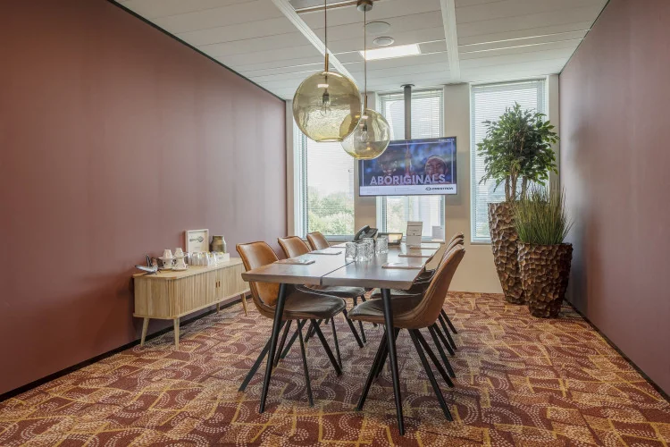 Vergaderruimte met houten tafel, lederen stoelen, grote planten en een tv-scherm aan de muur.